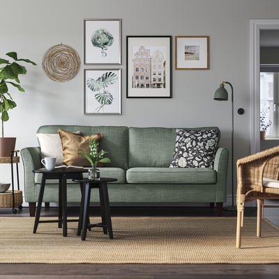 Moderne woonkamer met EKHOLMA bank, groen, pluche, 3-zits. Kunst aan de muur, vloerlamp, rieten stoel en vloerkleed. Strak, minimalistisch ontwerp.