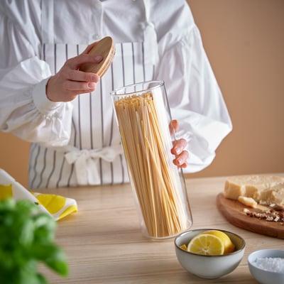 Een doorzichtige glazen pot genaamd EKLATANT met een houten deksel. Het is cilindrisch, geschikt voor het bewaren van voedsel en heeft een eenvoudig, elegant ontwerp.