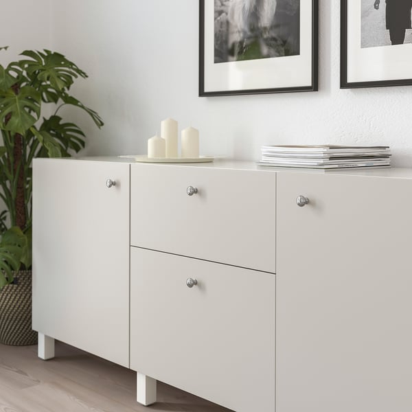 Witte kast met twee lades en deuren, chromen handgrepen, bekroond met kaarsen en boeken, naast een grote groene plant.