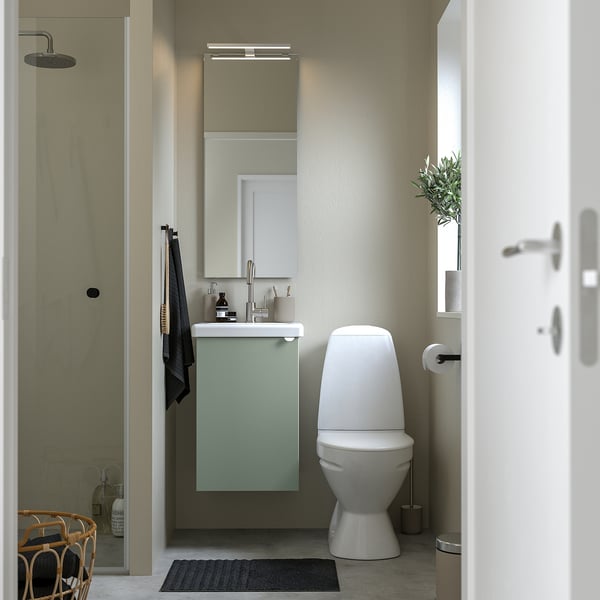 Moderne badkamer met licht grijsgroene ENHET wastafelkast, minimaal toilet, wandwastafel, verlichte spiegel en zwart vloerkleed.