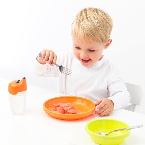 Het kind eet van sinaasappelkom die roestvrij staalvork gebruikt. Vaatwasmachinebestendig, leeftijd 3+. Helder, leuk, kindvriendelijk.