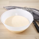 Witte kom met schuimige vloeistof, metalen garde, gestreepte doek op aanrecht.