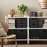 Moderne boekenkast met FJÄDERHARV donkere dozen voor papieren en boeken, elegante vaas met bloemen, stoelen, lichte houten vloer.