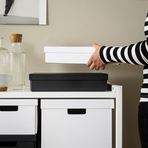 De hand reikt naar de witte FJÄDERHARV-doos bovenop het bureau, naast de zwarte doos en de karaf, met de nadruk op de grootte van de dozen en het moderne design.