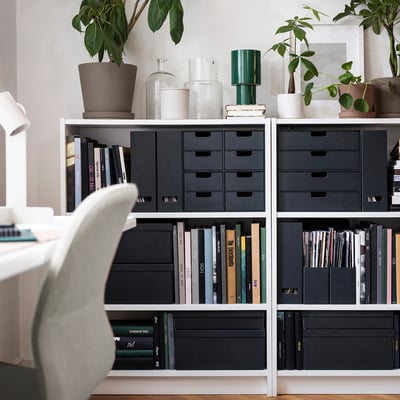 Moderne boekenkast met zwarte tijdschriften, boeken, planten en vazen. Stoel gedeeltelijk zichtbaar.