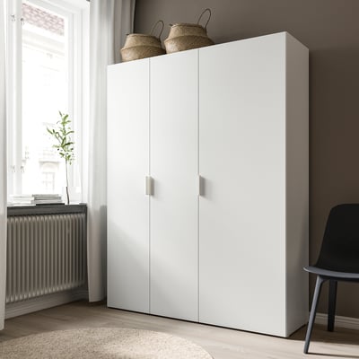 Witte platsa kledingkast met scharnierende deuren, twee rieten manden erop, bij raam met plant.
