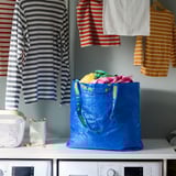 Blauwe herbruikbare boodschappentas, gevuld met kleurrijke kleding, zittend in de buurt van gestreepte shirts op hangers en twee wasmachines.