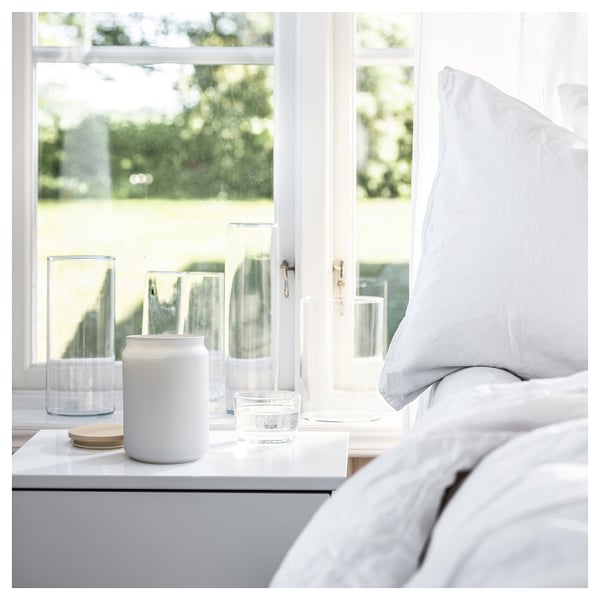 Schone slaapkamer bij raam, witte kaars, glas, houten onderzetter, heldere glazen beker op grijs nachtkastje.