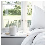 Schone slaapkamer bij raam, witte kaars, glas, houten onderzetter, heldere glazen beker op grijs nachtkastje.