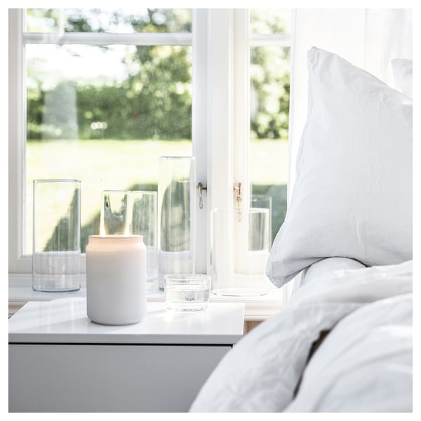 Een gezellige slaapkamerscène. Een witte kaars zit op een vensterbank naast heldere glazen, met een grijs bed zichtbaar.