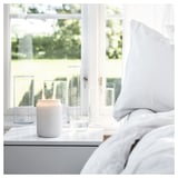 Een gezellige slaapkamerscène. Een witte kaars zit op een vensterbank naast heldere glazen, met een grijs bed zichtbaar.