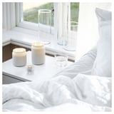 Heldere, witte slaapkamerscène met brandende kaarsen in de buurt van een raam. Strakke, minimalistische inrichting met fris beddengoed.