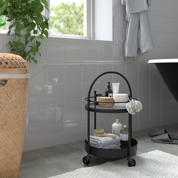 Kleine FRÖSJÖN zwart metalen badkamertrolley met glazen blad, met handdoeken, borstels en crèmes, naast een grote rieten wasmand en grijze tegels.