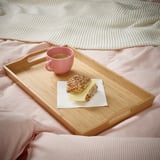 Houten ontbijtblad op een bed met roze lakens, met een kopje koffie en een broodje, voor een gezellige ontbijtervaring.
