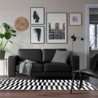 Moderne woonkamer met zwarte bank, grijze kussens, kunst, geruit tapijt, lamp, plant.