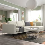 Moderne woonkamer met witte FULLERÖ bank, gezellige kussens, geometrische tafel en groene muren. Groot raam en levendig tapijt.