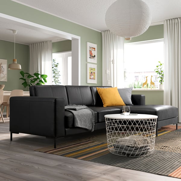 Moderne woonkamer met zwart lederen FULLERÖ bank, geel/grijze kussens, groene muren, groot raam, metalen salontafel en gestreept vloerkleed.