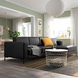 Moderne woonkamer met zwart lederen FULLERÖ bank, geel/grijze kussens, groene muren, groot raam, metalen salontafel en gestreept vloerkleed.