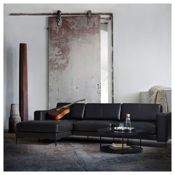 Moderne woonkamer met zwart lederen sectionals, ronde salontafel en grote rustieke schuifdeur. Gitaar, waterfles en boeken op tafel.
