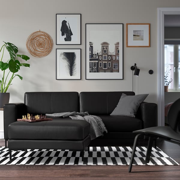 Moderne woonkamer met zwarte bank, grijze kussens, dambordkleed, galerijmuur, schaakspel, plant en vloerlamp.