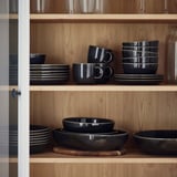 Houten planken met donkergrijze GLADELIG aardewerken kommen en mokken, netjes gestapeld, met heldere glazen erboven.