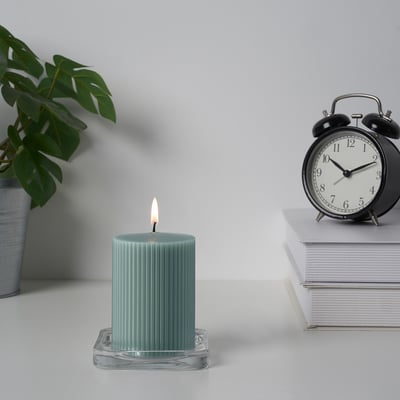 Een brandende groene kaars zit op een glazen houder naast een wekker en een stapel boeken.