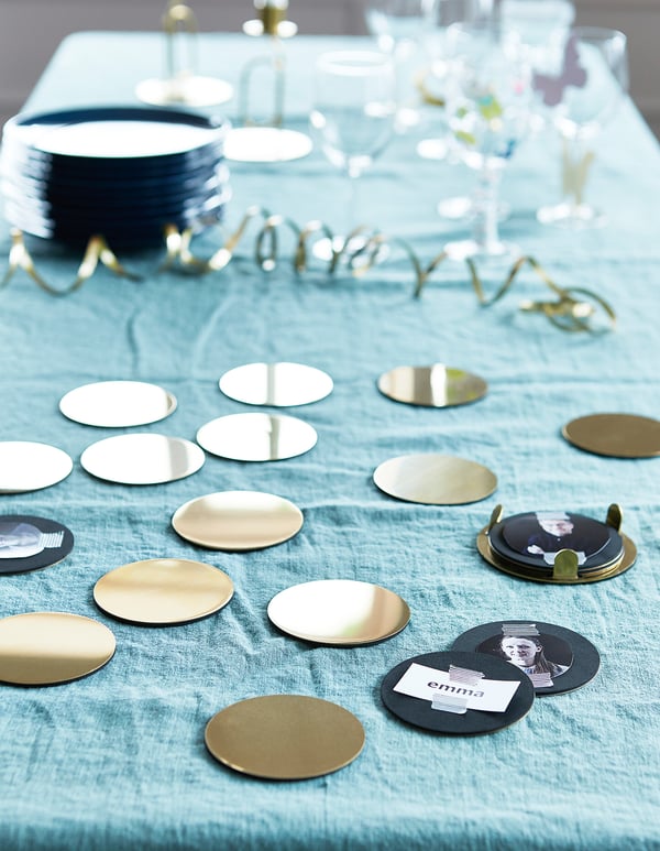 Tafel gedekt met blauwe stof, verspreide metalen onderzetters, sommige met namen. Linksboven stapel marineblauwe onderzetters op stand. Elegant, reflecterend ontwerp.