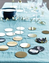 Tafel gedekt met blauwe stof, verspreide metalen onderzetters, sommige met namen. Linksboven stapel marineblauwe onderzetters op stand. Elegant, reflecterend ontwerp.