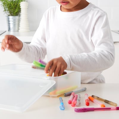 Kind dat kleurrijke pennen en tellers in een duidelijke stapelbare plastic doos op een witte lijst organiseert.