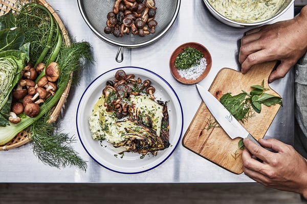 Chef-kok die maaltijd bereidt met verse champignons, venkel en kruiden. Plaat van voedsel, snijplank met mes, kommen en groenten.