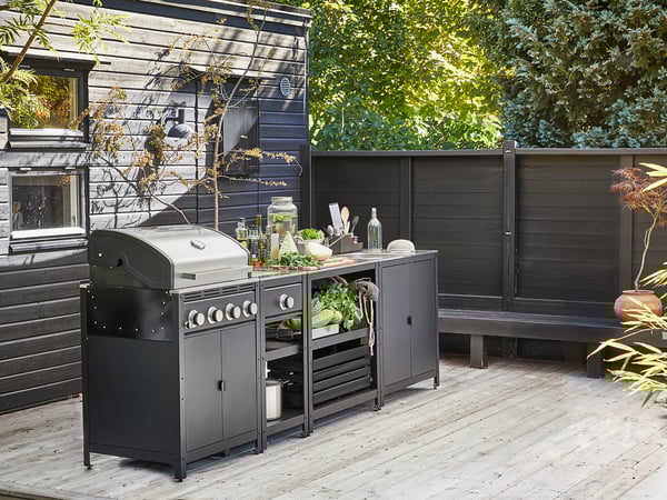 Buitenkeukenset met zwarte grill, gootsteen en planken op een houten terras, omgeven door donkere muren.