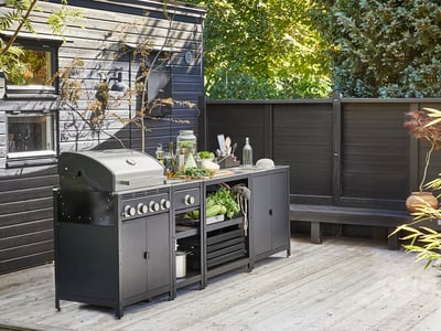 Buitenkeukenset met zwarte grill, gootsteen en planken op een houten terras, omgeven door donkere muren.