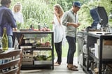Vier socializen rond het stalen kookeiland van GRILLSKÄR met groenten en grillgereedschap. Heeft gegalvaniseerde planken om buiten te koken.