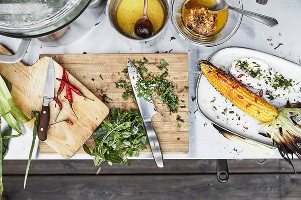 GRILLSKÄR kookeiland, zilver, roestvrijstalen blad, rechthoekig, chromen handgrepen, twee houten planken, geprepareerde grillmaïs, messen, kruiden.
