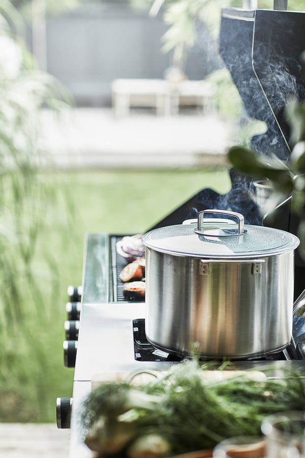 Een zilveren gasgrill voor buiten met deksel, geplaatst op een groene veranda, met kookroosters en knoppen zichtbaar.