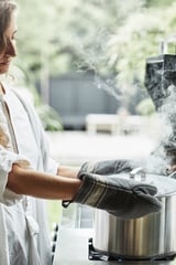 Vrouw die buiten kookt met behulp van witte GRILLSKÄR-grill met opstijgende rook, met grijze ovenwanten, tegen een groene tuinachtergrond.