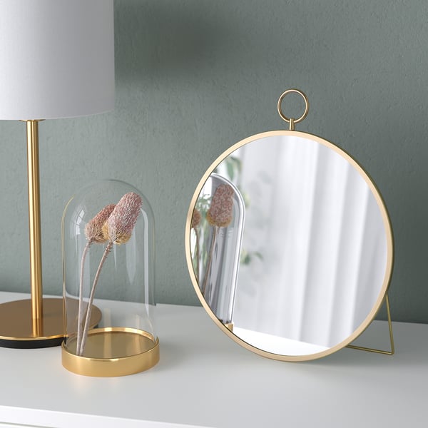 Een moderne spiegel met een gouden standaard en lus aan de bovenkant, die een lamp en muur weerspiegelt. Het kan worden opgehangen of vrijstaand worden gebruikt, uitgestald op een witte tafel.