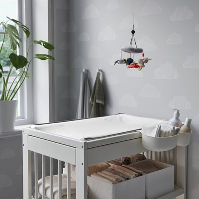 Kinderkamer met witte GULLIVER commode, knuffels, luiers en grijs wolkenbehang. Plant en handdoeken in de buurt.