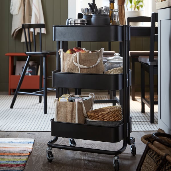 Zwarte metalen trolley met 3 niveaus en gevlochten manden in de keuken. Bevat fruit, kruiden en keukengerei.