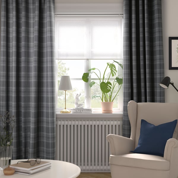 Moderne woonkamer met HÄGGVECKMAL donkergrijze tartan kamer-verduisterende gordijnen, witte jaloezieën, gezellige stoel, lamp en potplant op een witte vensterbank.