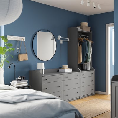Moderne blauwe slaapkamer met HAUGA grijze lades en kledingkast, ronde spiegel, witte lamp, planten en koptelefoon op boeken.