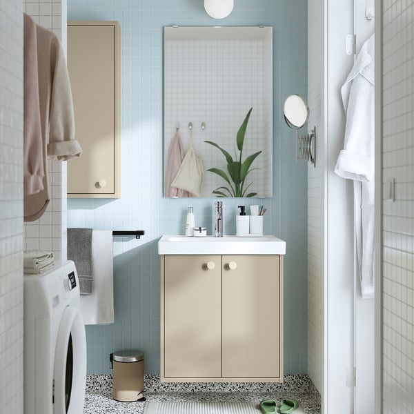 Klein, beige kastje in de moderne badkamer; Lichtblauwe tegels, witte wastafel en wasmachine, spiegel, hangende handdoeken.