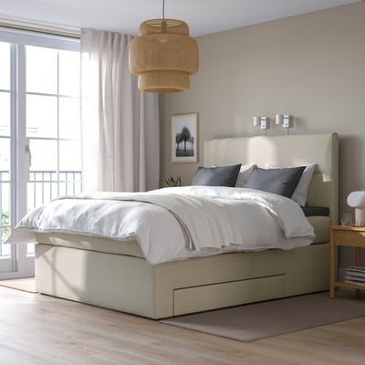 Moderne slaapkamer met groot beige platformbed, wit beddengoed, grijze kussens en natuurlijke hanglamp.