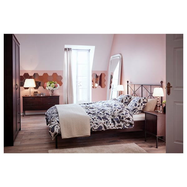 Gezellige slaapkamer met roze muren, zwart metalen bed, beddengoed met patroon, houten lades en grote vloerspiegel.