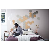 Een persoon zit op een bed met witte dekens tegen een muur versierd met zeshoekige spiegels in grijs en bruin, waardoor een moderne, rustige slaapkamerscène ontstaat.