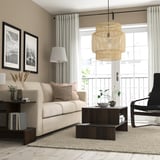 Minimalistische woonkamer met beige bank, houten salontafel, zwarte fauteuil, groot raam en hanglamp. Beige muren en neutraal vloerkleed.