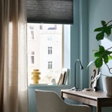 Thuiskantoor met blauwe muren, grijze jaloezieën, houten bureau, lamp, stoel, plant en gele vaas.