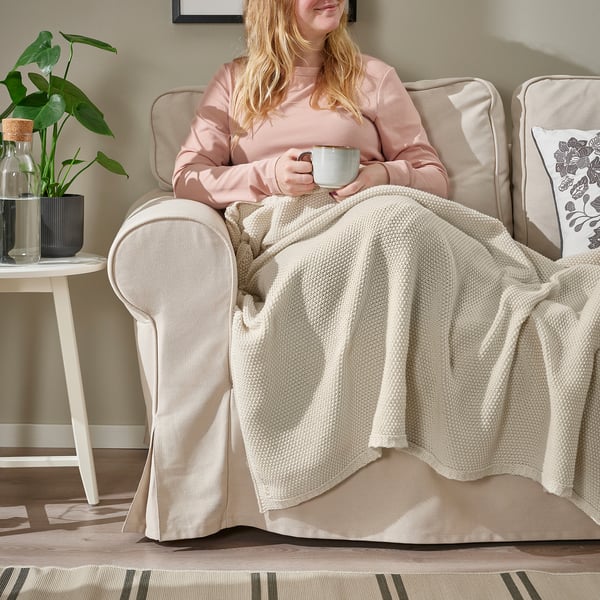 Vrouwenzitting op beige stoel, deken over benen, houdende koffiekopje.