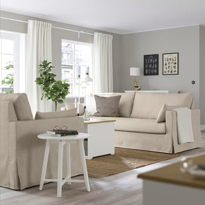 Moderne woonkamer met HYLTARP beige bank, fotolijsten, wit vloerkleed, bijzettafel en lamp.