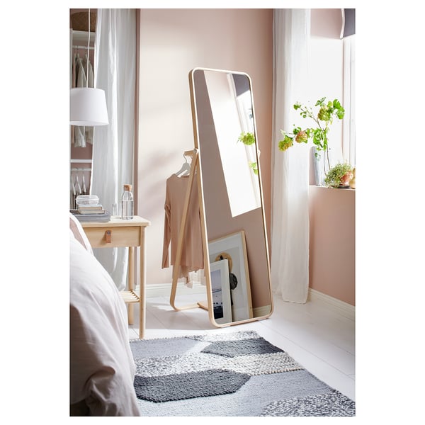 Slaapkamer met wit bed, houten tafel en goudomlijste vloerspiegel die een kledingrek en raam weerspiegelt.